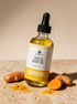 Kokoualush Turmeric Face & Body Oil in a 2oz glass dropper bottle on travertine with fresh turmeric root and golden powder