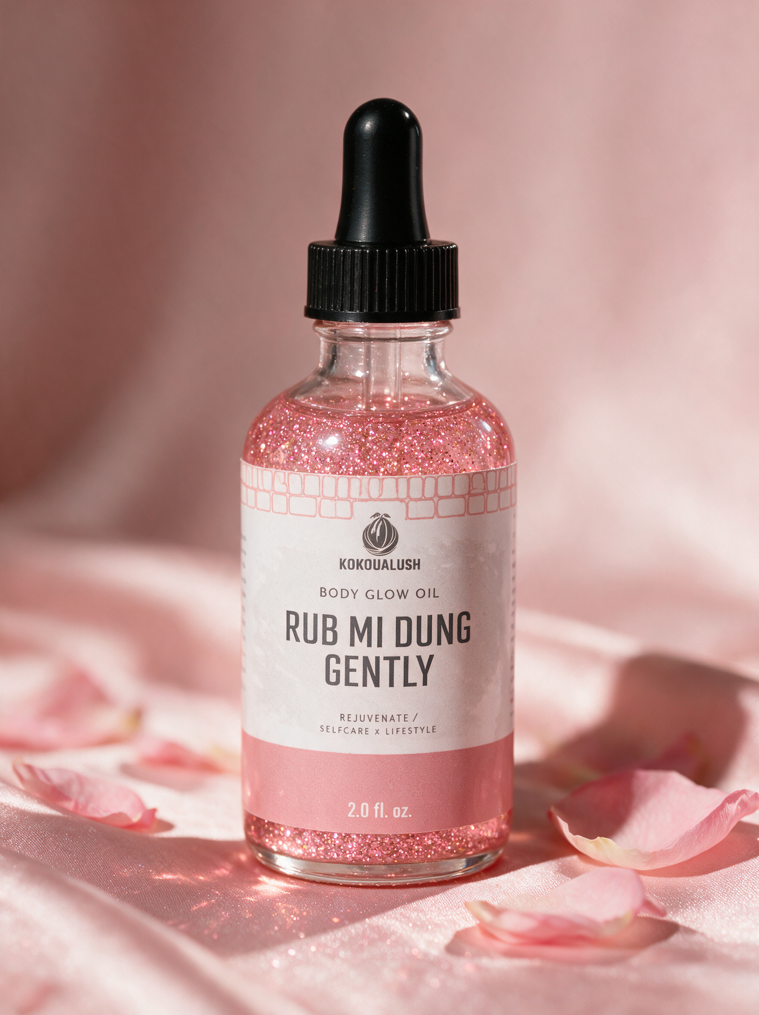 Kokoualush Rub Mi Dung Gently pink shimmer Body Glow Oil dropper bottle on blush silk with rose petals