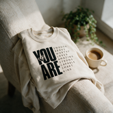 You Are Enough sand crewneck draped over a linen armchair in warm light